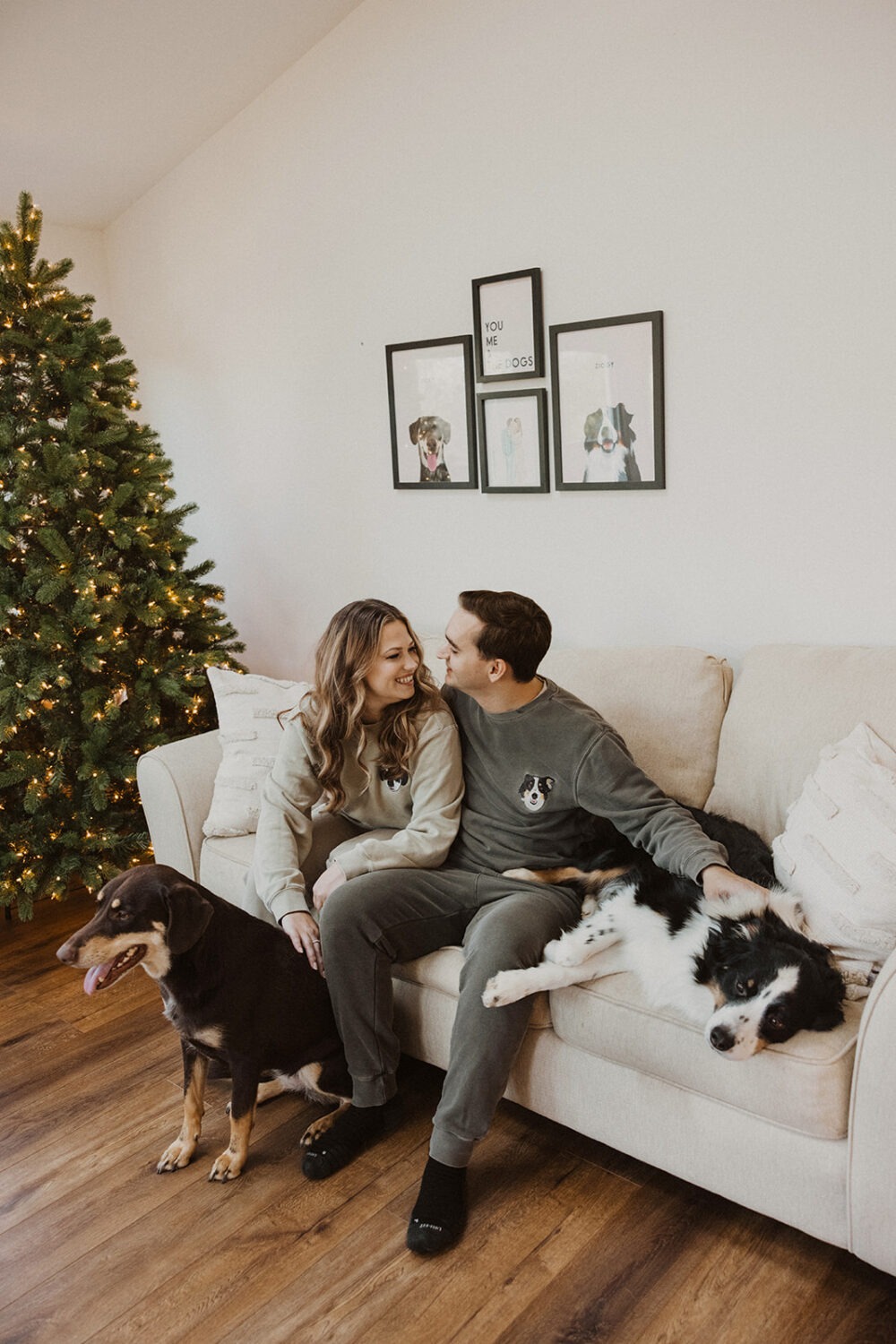 couple sits with dogs in front of Christmas tree at winter engagement photoshoot