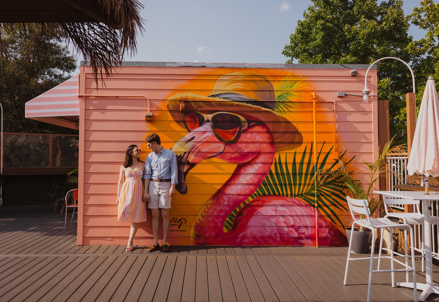 couple holding hands in front of pink flamingo wall during engagement shoot