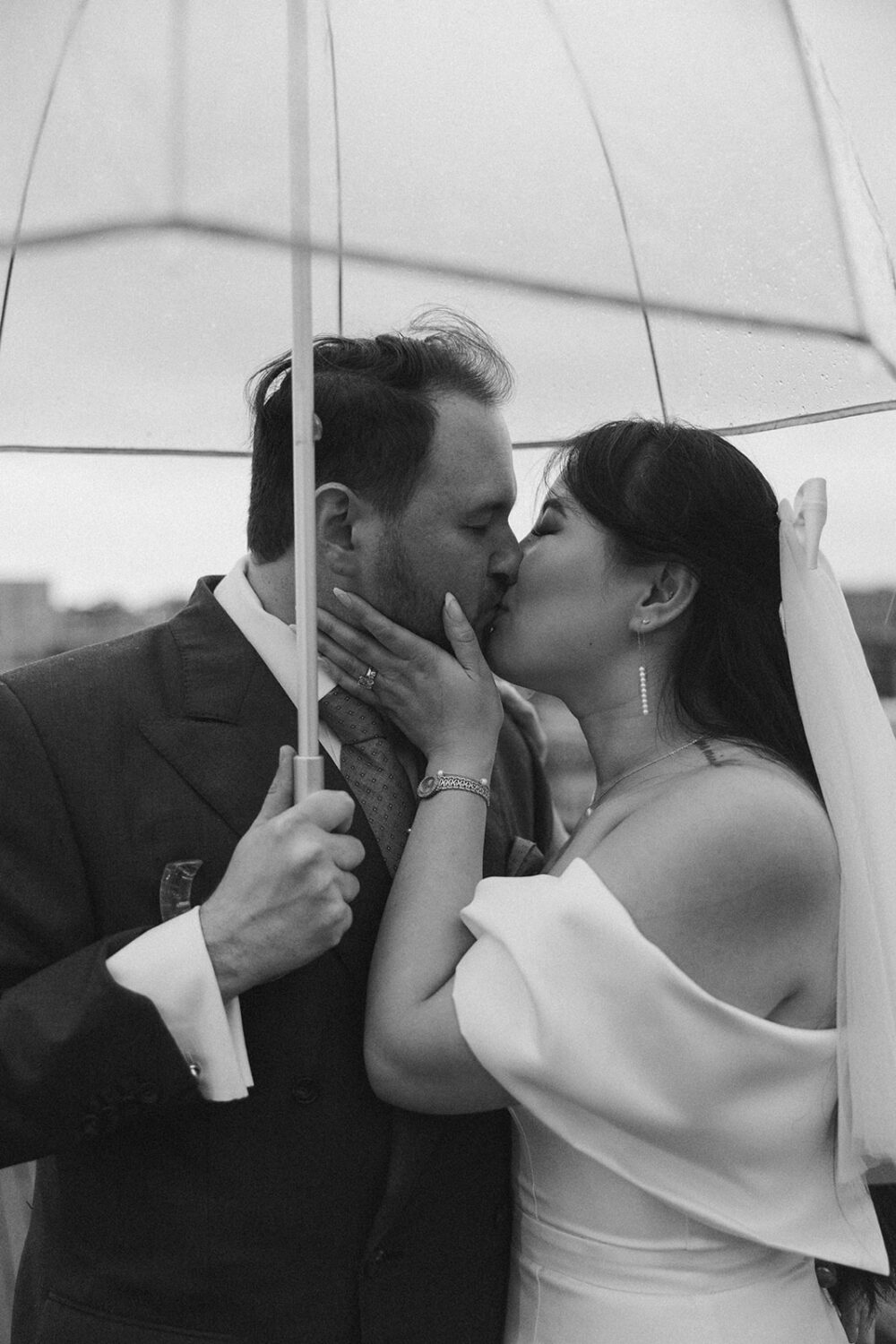 couple kiss while holding clear umbrellas during rainy wedding day