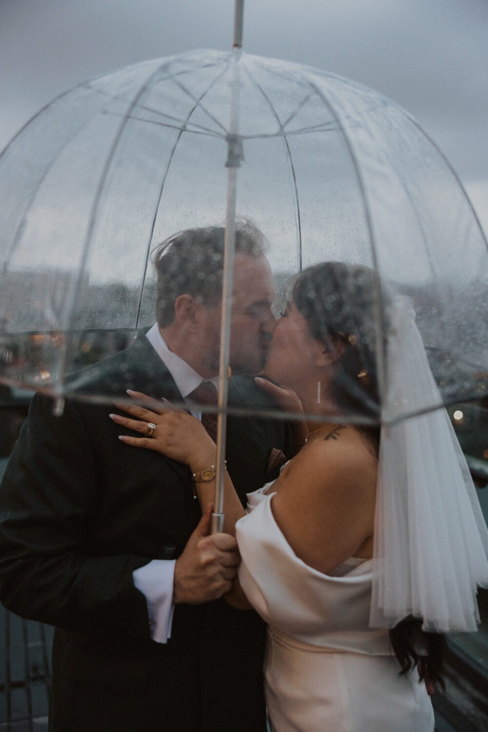 couple kiss while holding clear umbrellas during rainy wedding day