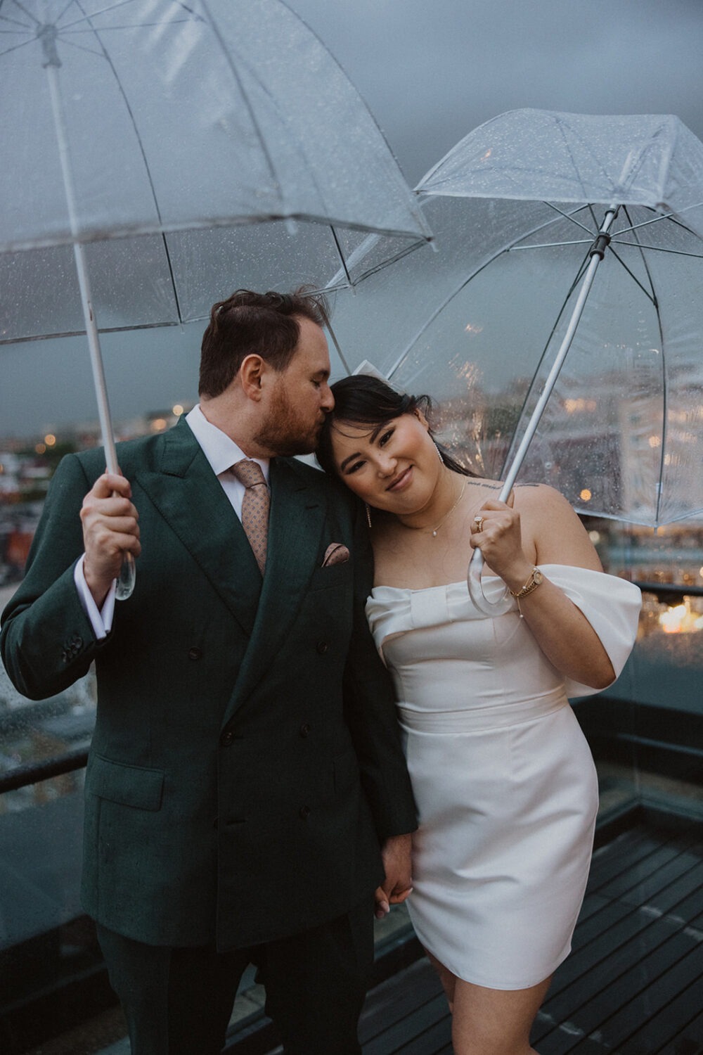 couple embrace while holding clear umbrellas during rainy wedding day