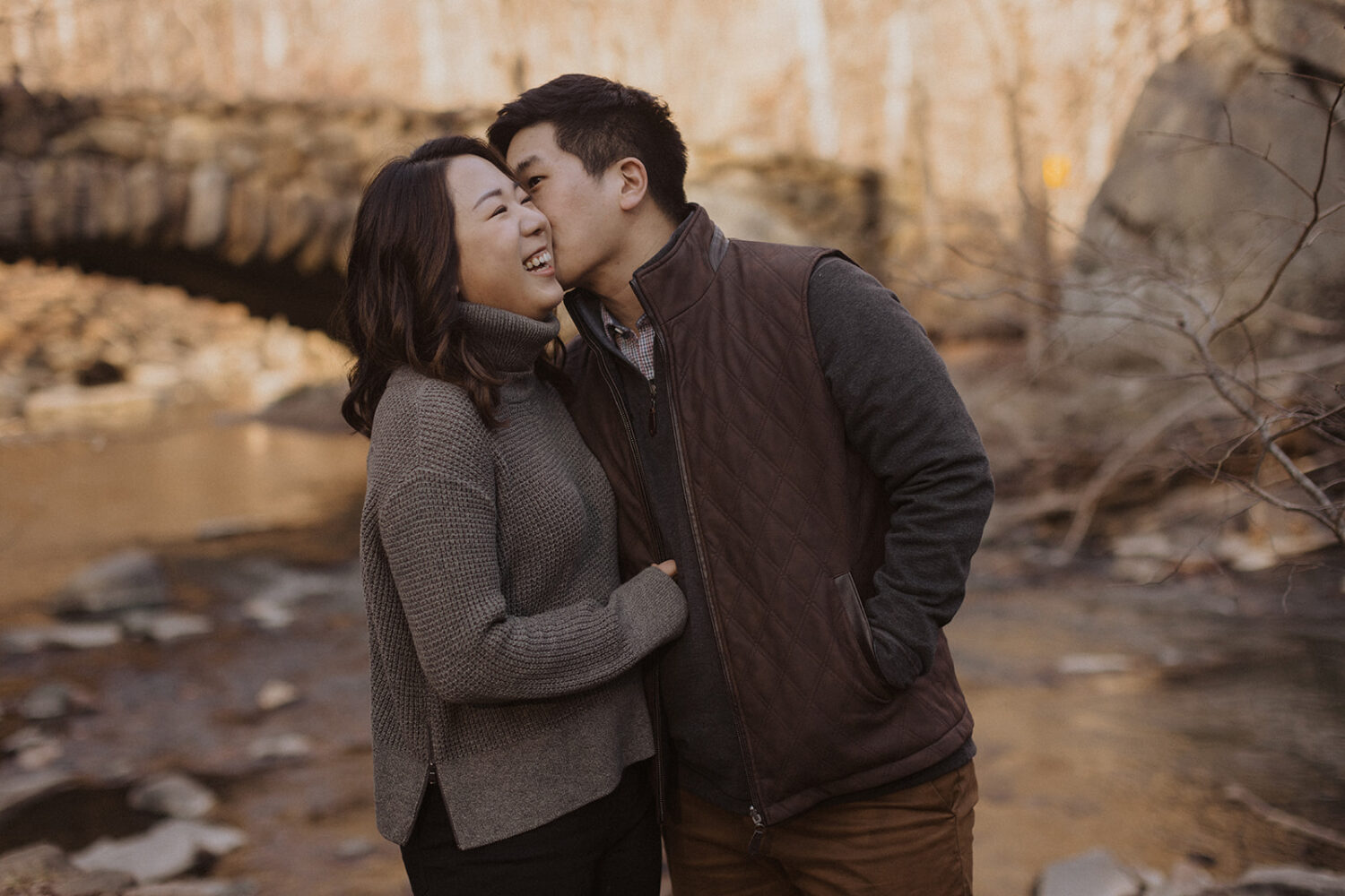 couple kiss while taking winter engagement photos by bridge