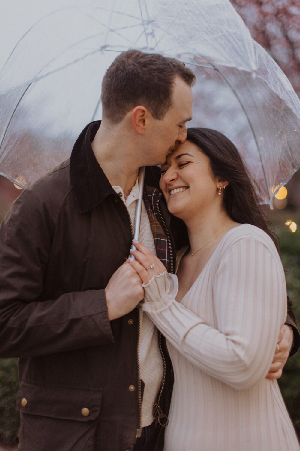 couple embrace under clear umbrella with pink flowering trees around them