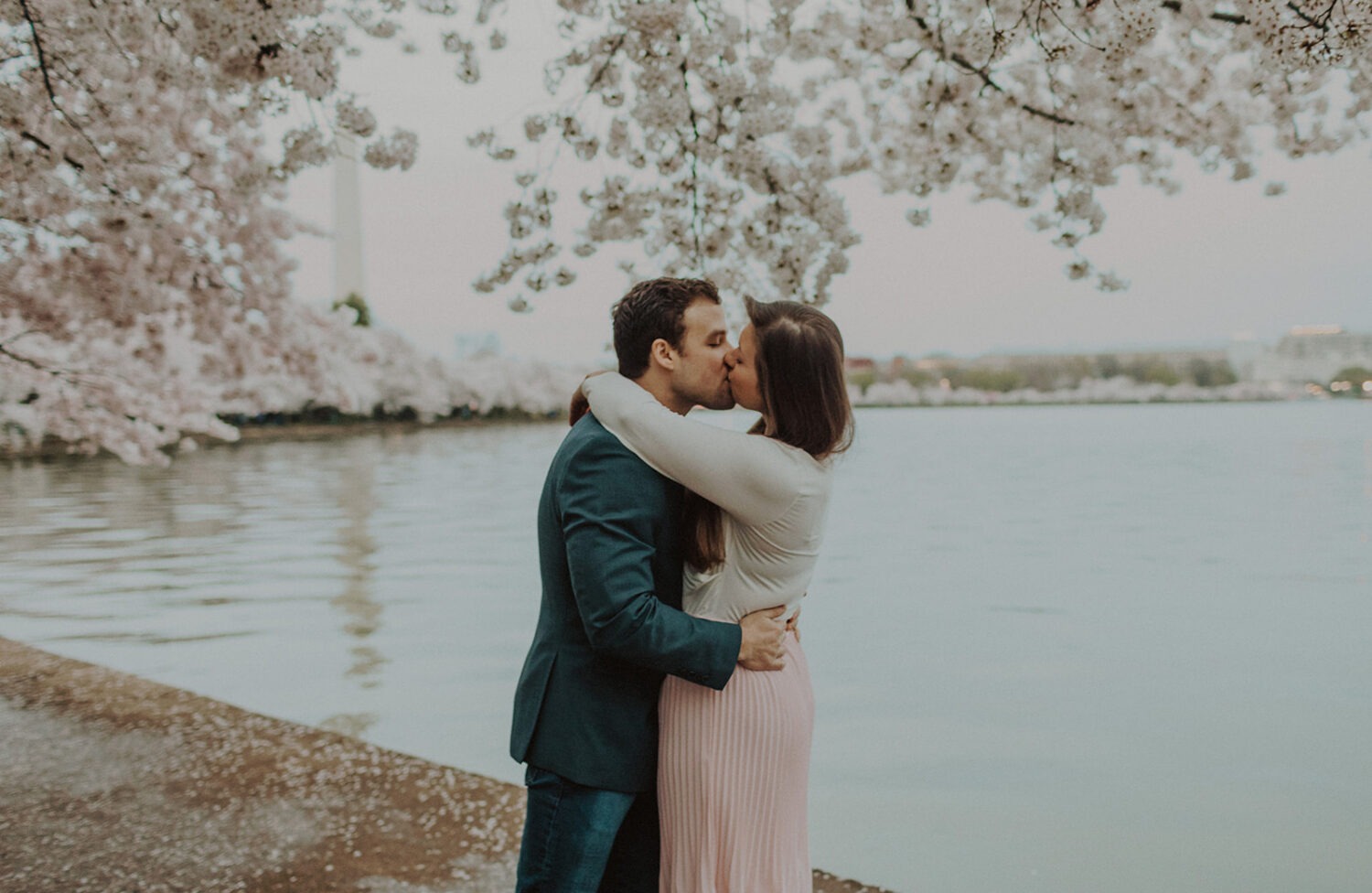 couple kiss on ledge along the water surrounded by cherry blossom trees