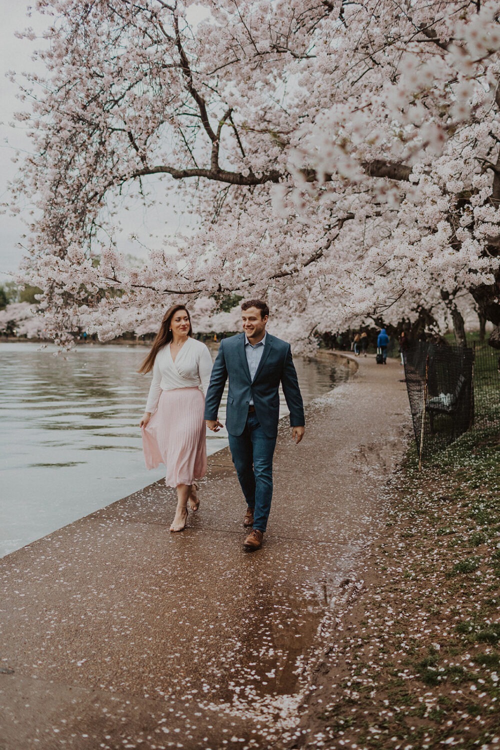 couple walk together holding hands along the water while taking DC cherry blossom photos