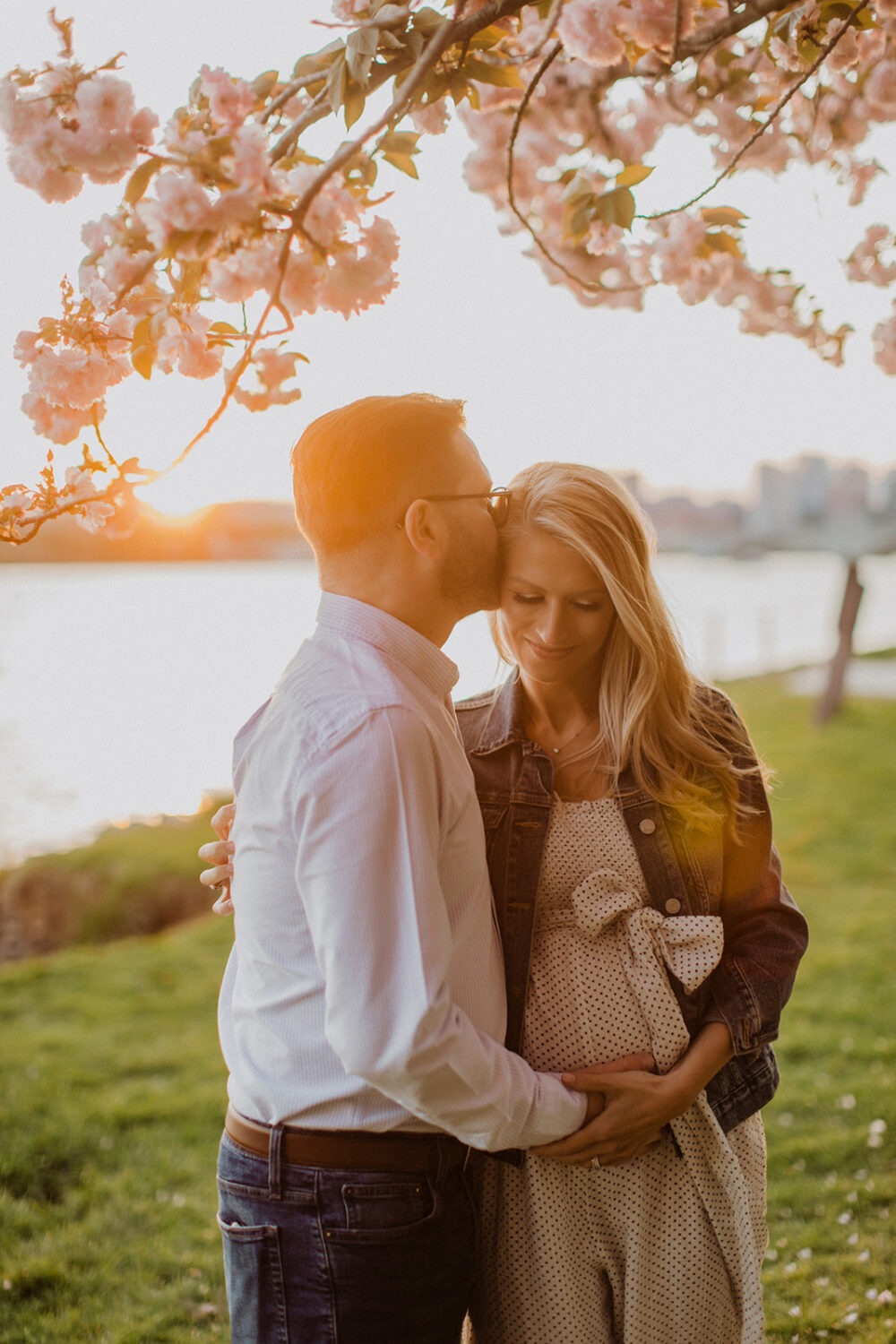 couple pose while taking maternity photos in the middle of cherry blossom trees
