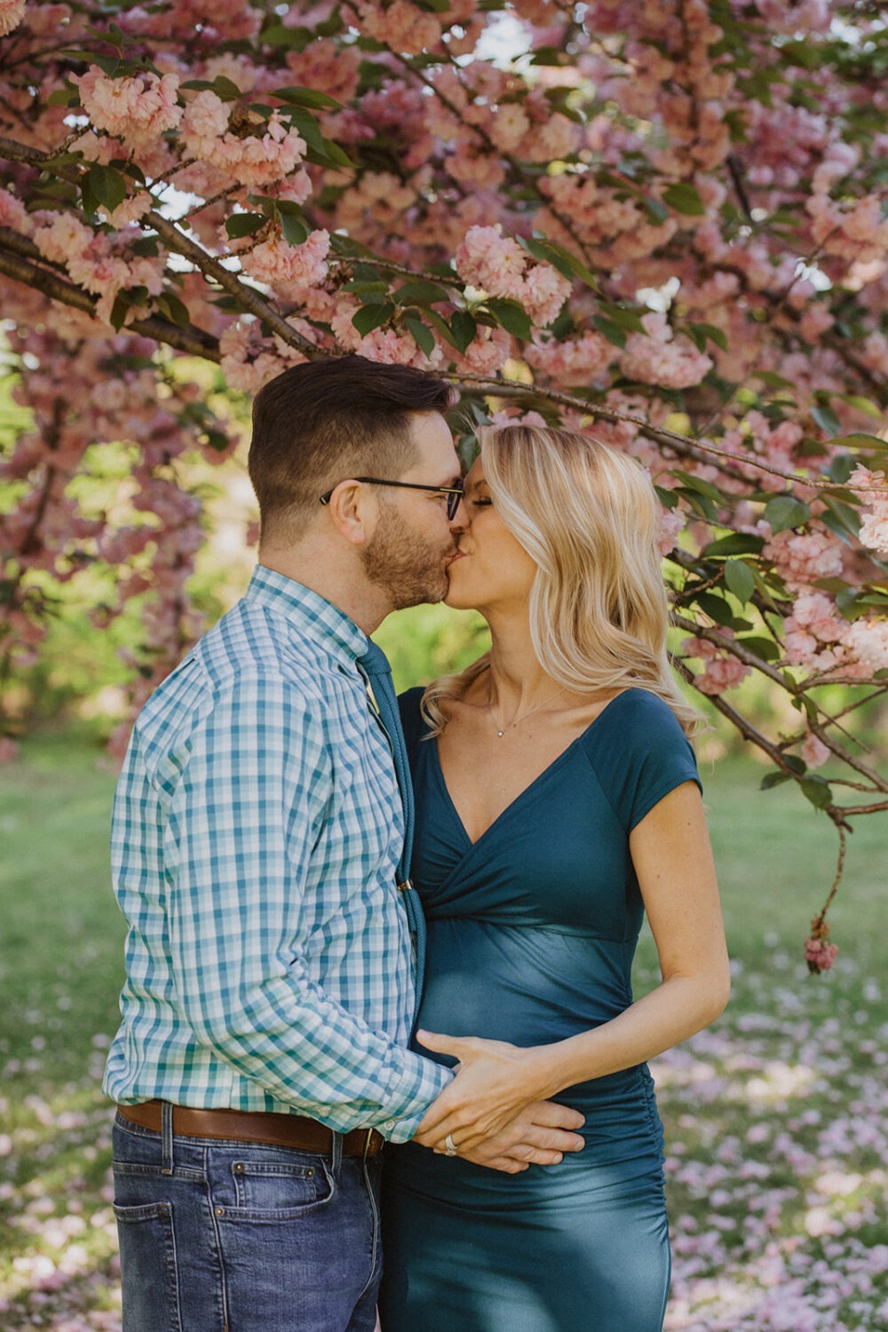 couple kiss while taking maternity photos in the middle of cherry blossom trees