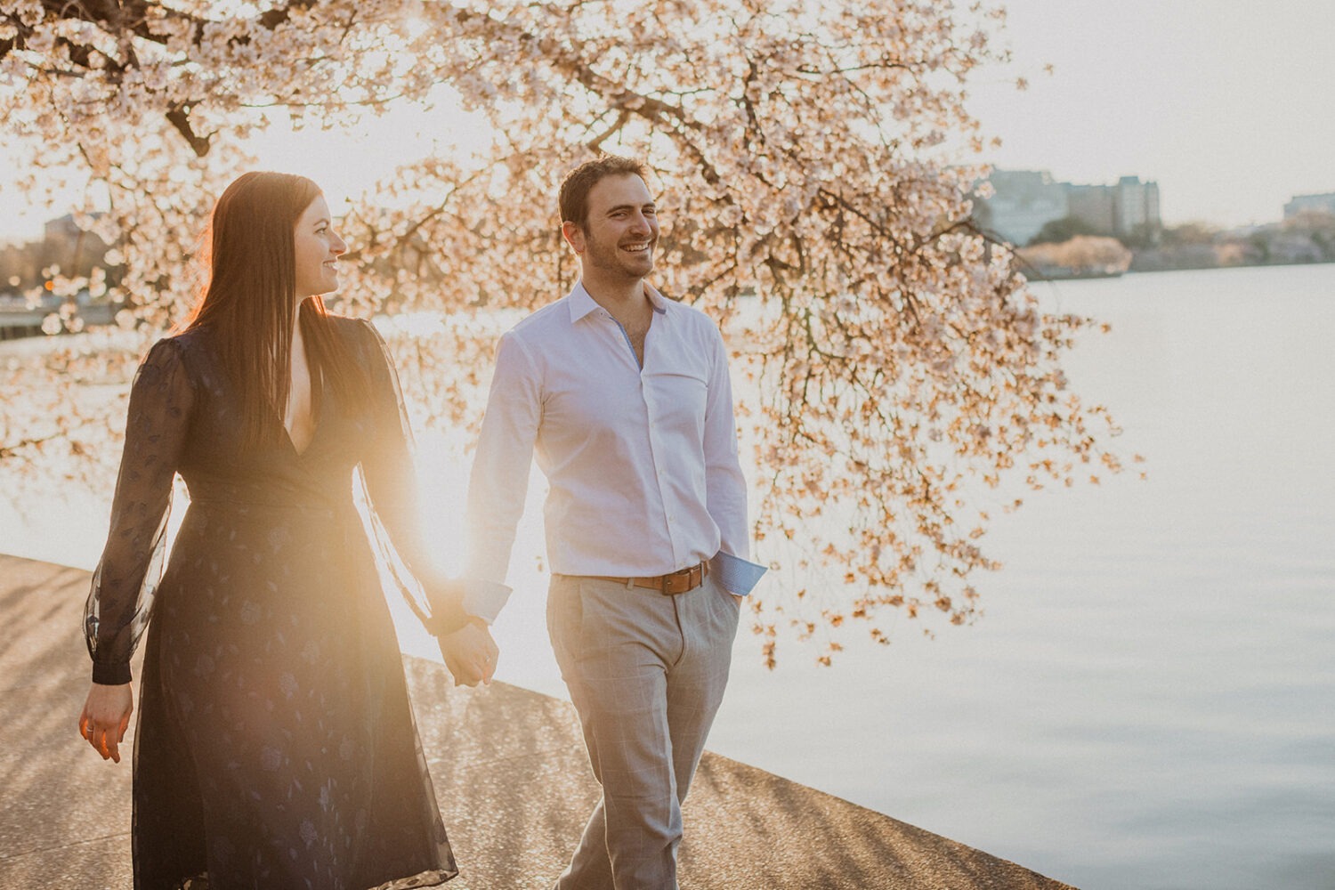 couple walk together holding hands along the water while taking DC cherry blossom photos at sunset