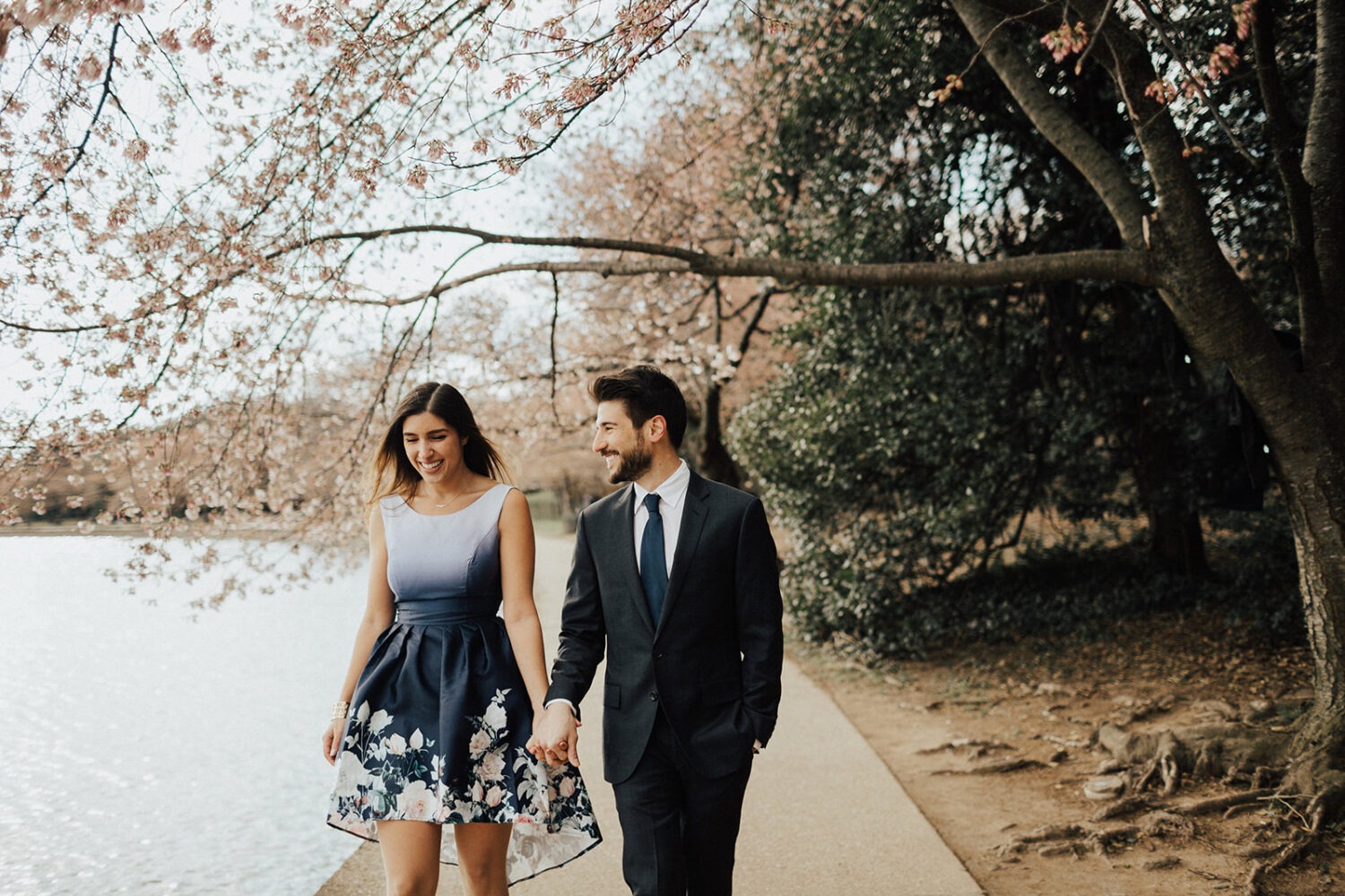 couple walk together holding hands along the water while taking DC cherry blossom photos