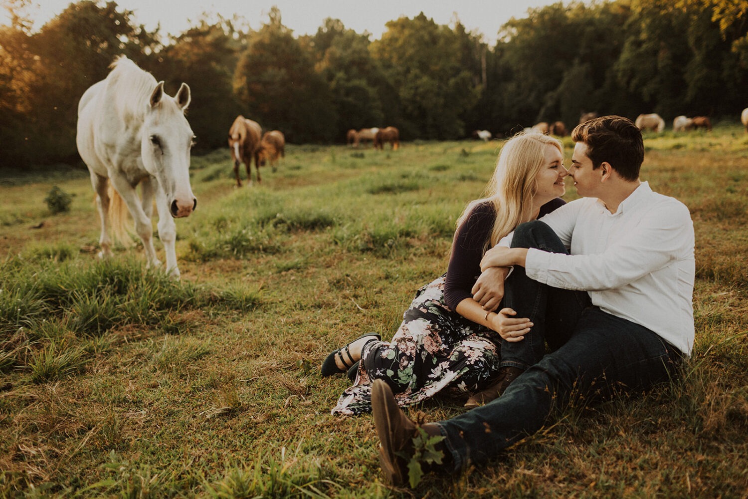 couple pose together while sitting in field surrounded by horses