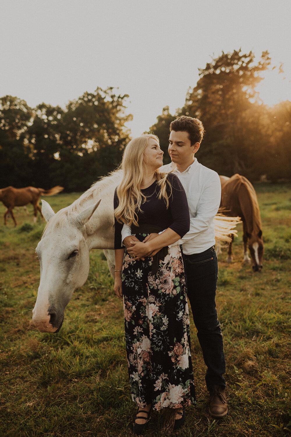 couple pose in a field of horses at sunset
