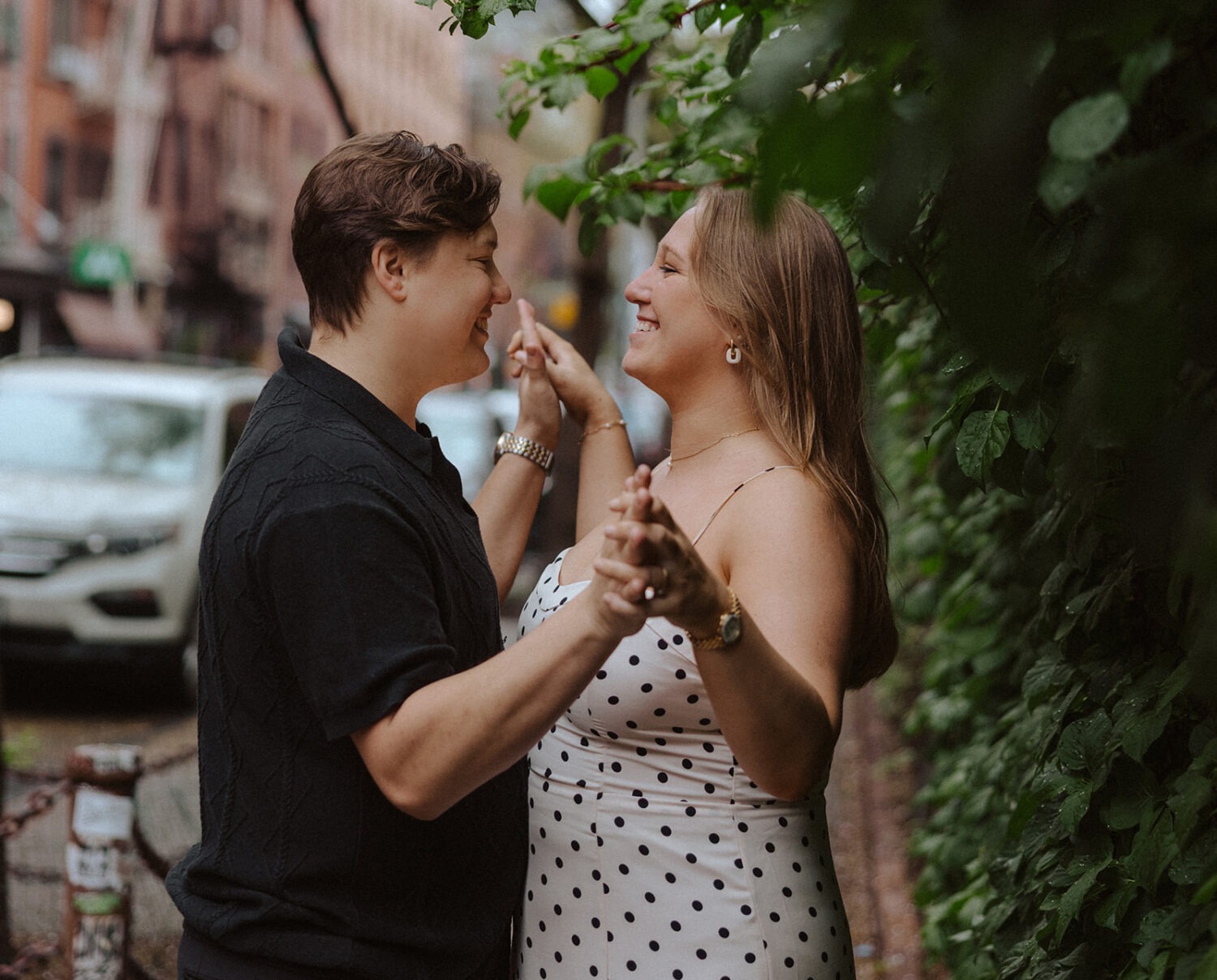 couple hold hands looking at each other against bush during rainy engagement photos