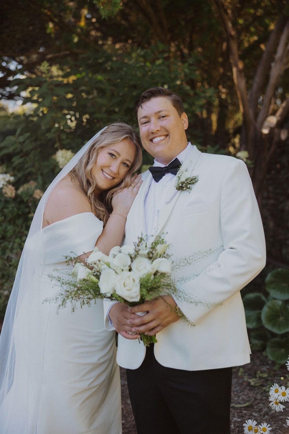 couple pose together as they both wear white and have white floral bouquet